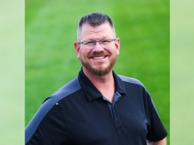 Corporate headshot of a man in a black polo with a green grass background
