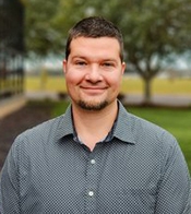 Corporate portrait of a man in a grey shirt with grass and trees in the background