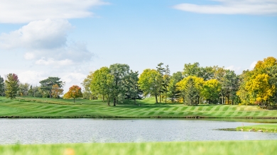 A green hill with lake in foreground and yellow and green trees in the distance with a blue sky.