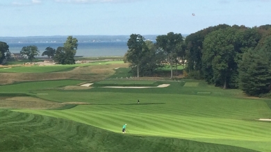 Overhead view of a green golf course with trees lining the side with ocean in the background
