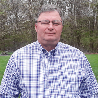 Headshot of a man in a checkered shirt with trees and grass in background