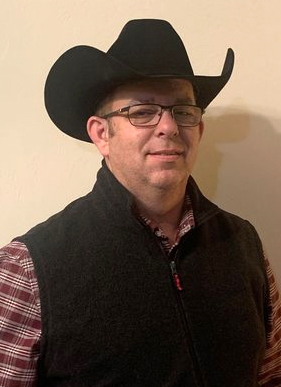Headshot of a man in a black cowboy hat and a black vest with red and white shirt. 