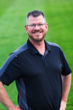 Corporate headshot of a man in a black polo with a green grass background