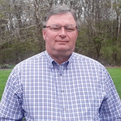 Headshot of a man in a checkered shirt with trees and grass in background