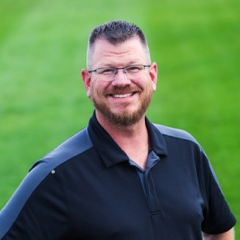 Corporate headshot of a man in a black polo with a green grass background